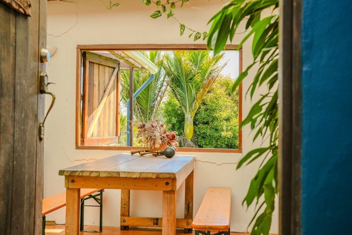 A wooden table with a bench, a window with a view of greenery, and a door with a rustic design.