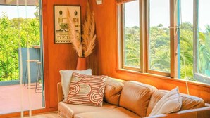 A cozy room with a wooden interior, a sofa with patterned cushions, a large window with a view of greenery, and a framed picture on the wall.