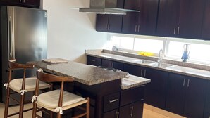 Uma cozinha moderna com armários escuros, geladeira de aço inoxidável, uma ilha central com banquetas e uma janela que traz luz natural.