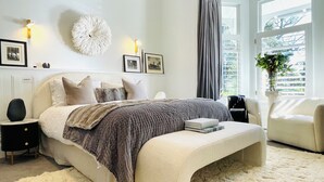A bedroom with a large bed, bedside tables, a bedside lamp, a wall-mounted fluffy decoration, framed pictures, a window with curtains, and a potted plant.