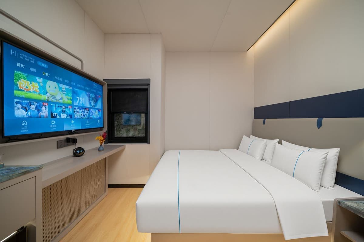 Une chambre d’hôtel moderne avec, à gauche, un grand téléviseur à écran plat, un lit avec une literie blanche et des touches de bleu, une petite fenêtre et une tête de lit bleu foncé.