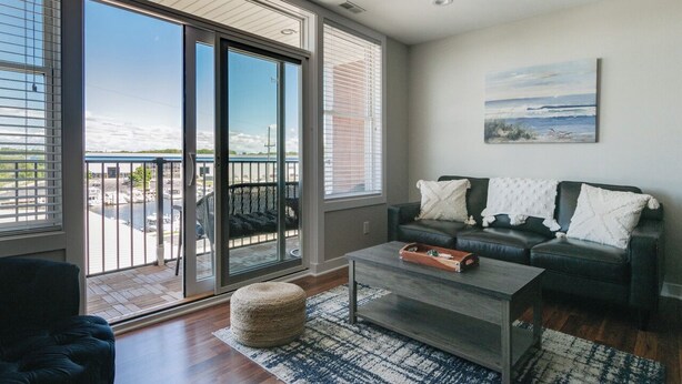 A modern living room with a large sliding glass door leading to a balcony, a dark leather sofa, a coffee table, and a painting on the wall. - Luxury Waterfront Condo (St. Joseph)
