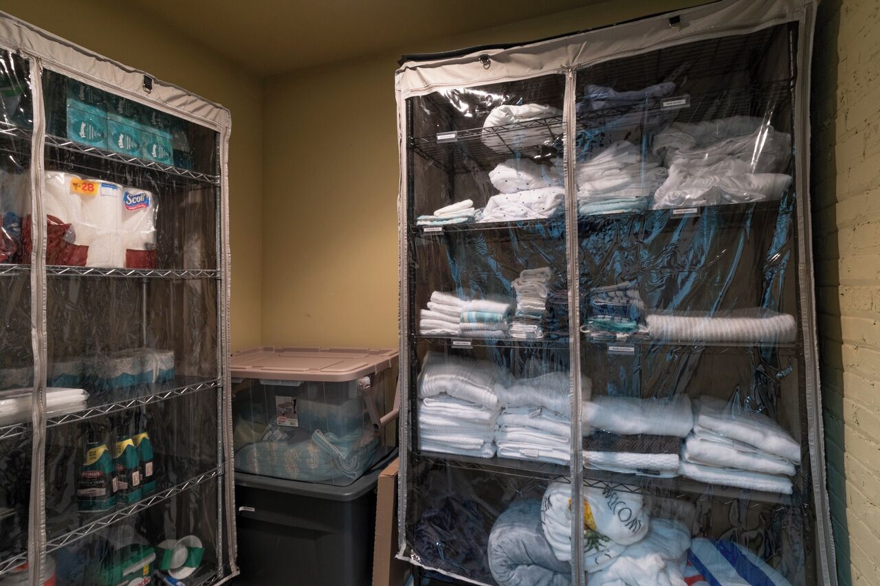 A storage room with shelves containing folded towels and linens, and a plastic bin with cleaning supplies.