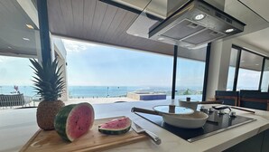 Una cocina moderna con una isla grande, tabla de cortar con piña y sandía, y vista al océano.