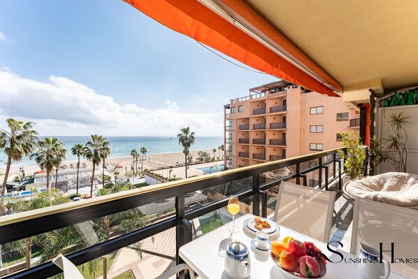 A balcony with a table set for breakfast, overlooking a beach and a pool.