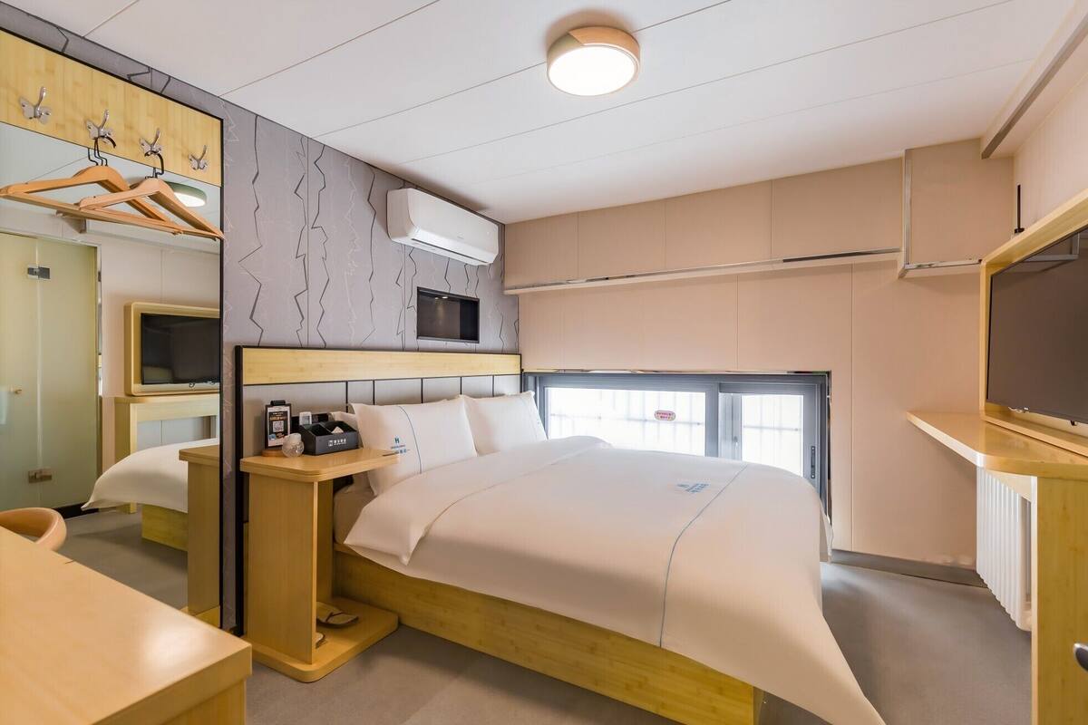 A modern hotel room with a bed, bedside table, TV, and a wardrobe with hanging clothes.