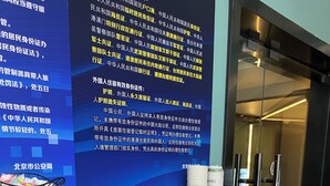 Zona de recepción de un hotel, con mostrador de recepción, una pared con carteles informativos y un ascensor de vidrio.