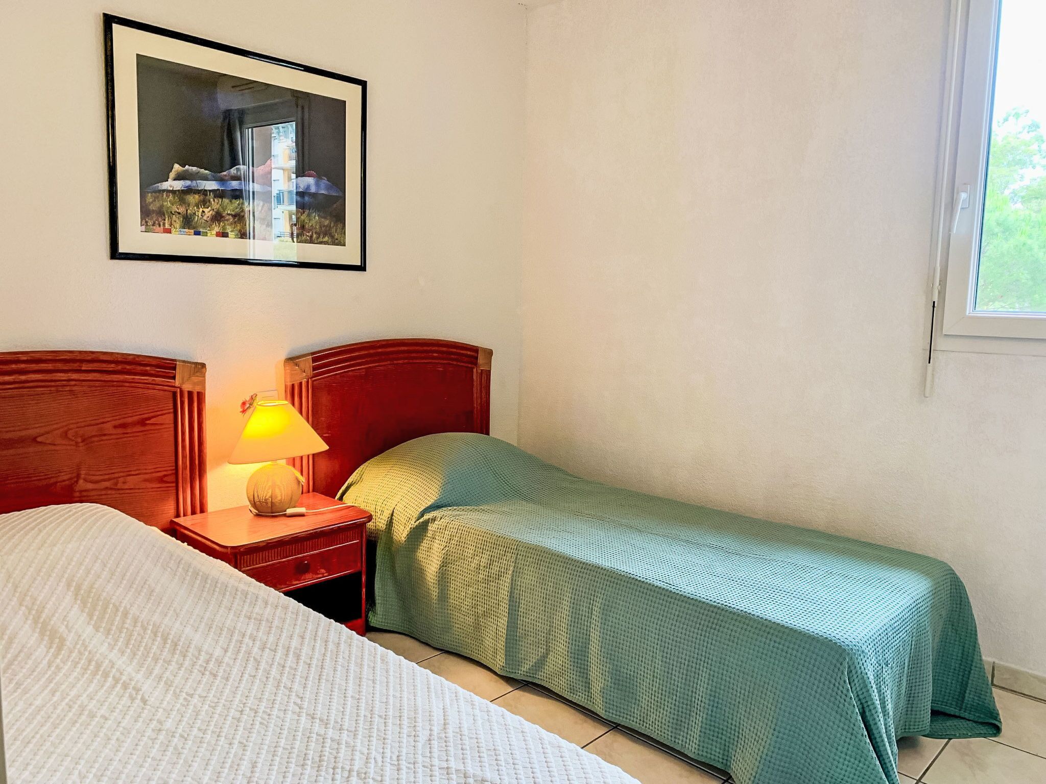 Une chambre avec deux lits, une table de chevet avec une lampe, un tableau encadré au mur et une fenêtre.