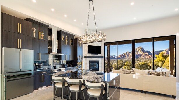 A modern kitchen with dark cabinetry, a central island with bar stools, a large window with a mountain view, and a cozy seating area with a sofa and cushions. - Zendona | Luxury Estate | Hot Tub, Putting Green and Fire Pits | Stunning Views | Near Trails (Sedona)