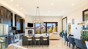 A modern kitchen with dark cabinetry, a central island with bar stools, a large window with a mountain view, and a dining area with a glass partition. - Zendona | Luxury Estate | Hot Tub, Putting Green and Fire Pits | Stunning Views | Near Trails (Sedona)