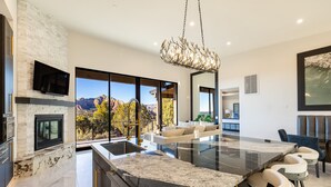 A modern kitchen with a large island, a flat-screen TV mounted on the wall, and a view of the mountains through large windows. - Zendona | Luxury Estate | Hot Tub, Putting Green and Fire Pits | Stunning Views | Near Trails (Sedona)