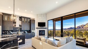 A modern kitchen with dark cabinets, a marble countertop, and a large island. There is a sofa with patterned cushions in the living area, and a large window offering a view of mountains. - Zendona | Luxury Estate | Hot Tub, Putting Green and Fire Pits | Stunning Views | Near Trails (Sedona)