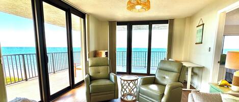 A spacious living room with a view of the ocean, featuring two green armchairs, a small round table, and a sofa.
