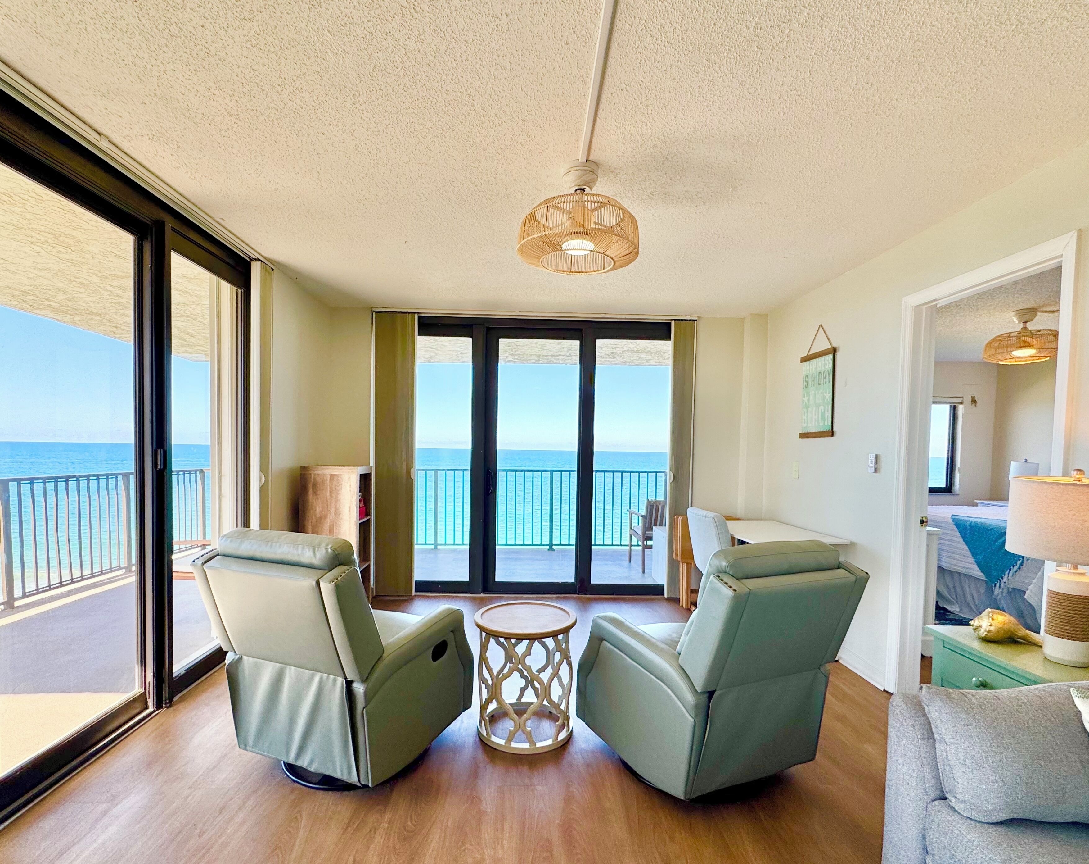 Een kamer met uitzicht op de oceaan, twee groene fauteuils, een kleine ronde tafel en een plafondlamp.