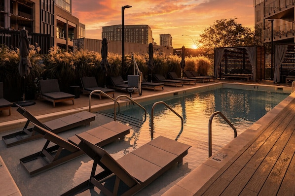 A rooftop pool with sun loungers and a view of the city skyline at sunset.