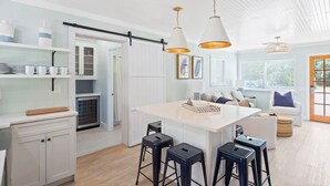 Cocina moderna con una isla central, taburetes de bar, luces colgantes y un comedor con sofá y cojines.
