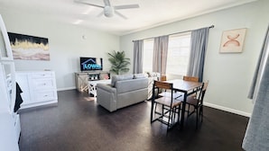 Un salon spacieux avec un canapé blanc, une table à manger avec des chaises, un ventilateur de plafond, un téléviseur à écran plat et une grande fenêtre avec des rideaux.