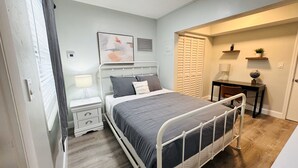 Une chambre avec un lit en métal, une table de chevet avec une lampe, un climatiseur fixé au mur et un petit bureau avec une chaise.