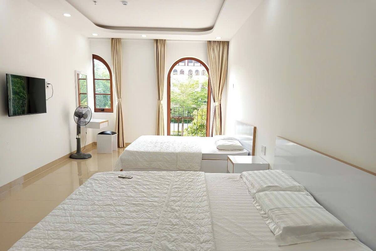A hotel room with two beds, a flat-screen TV, a bedside table, a fan, and a window with curtains.