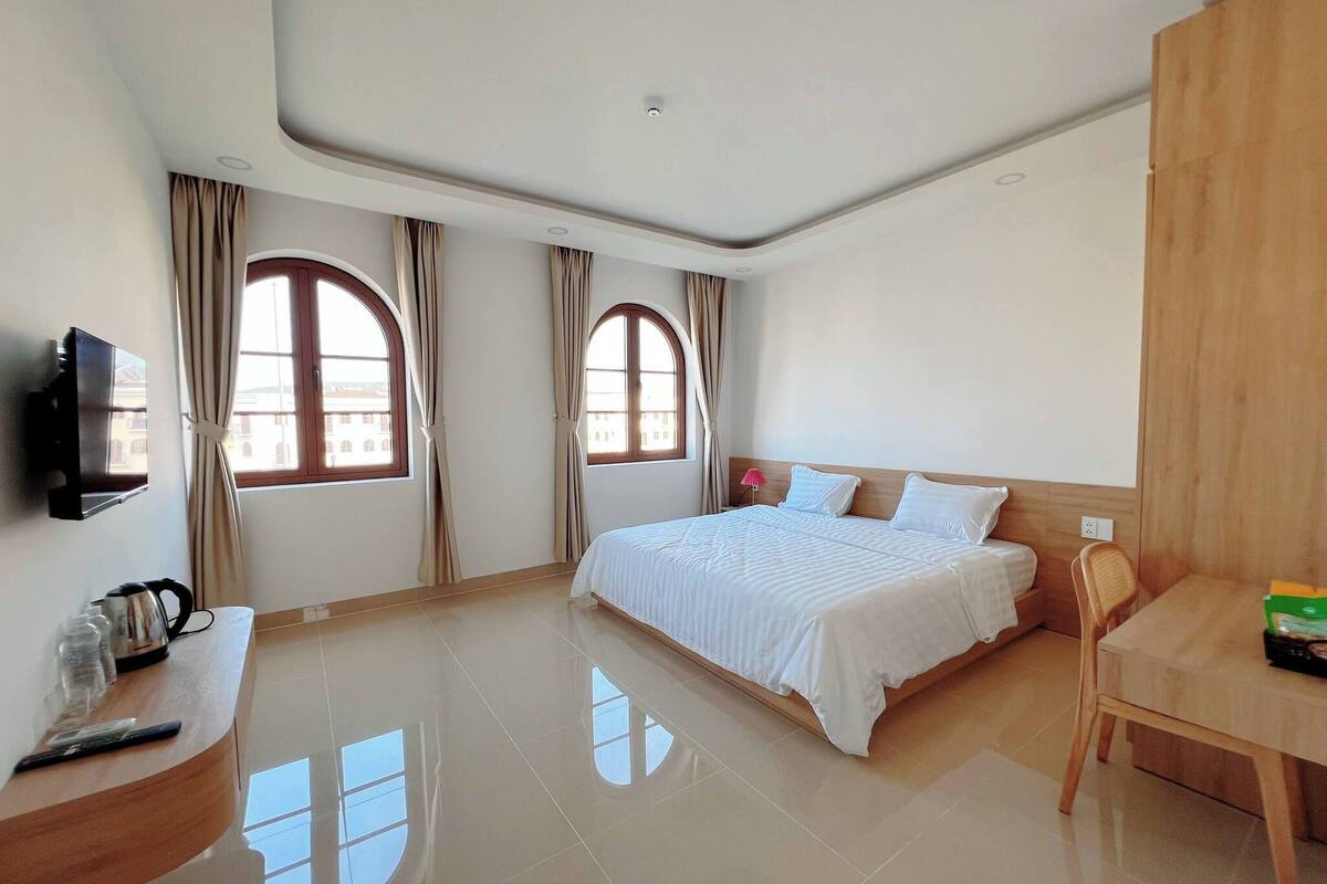 A hotel room with a large bed, two windows with curtains, a flat-screen TV, a wooden desk, and a chair.