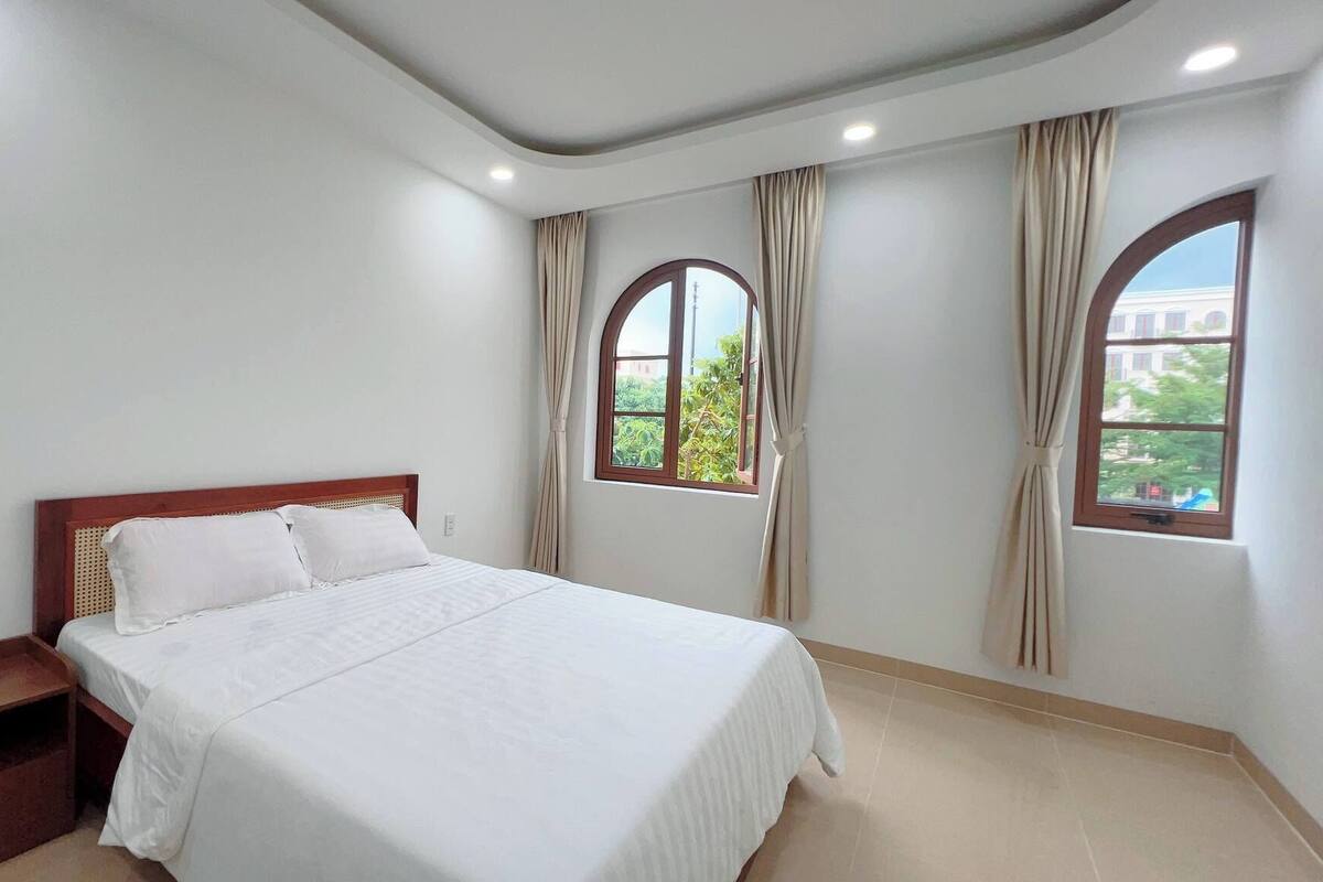 A bedroom with a bed, a nightstand, and two windows with curtains.