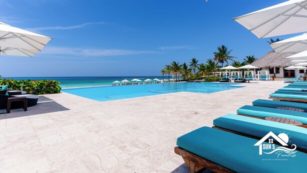 A resort pool area with lounge chairs and umbrellas, a clear blue sky, and a view of the ocean. - Refugio Ideal Golf & Piscina Cana Rock Star (Bavaro)