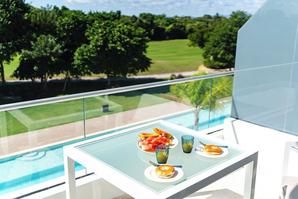 Une table à plateau en verre, sur laquelle sont disposés des aliments pour le petit-déjeuner, avec vue sur une piscine et sur la verdure environnante.