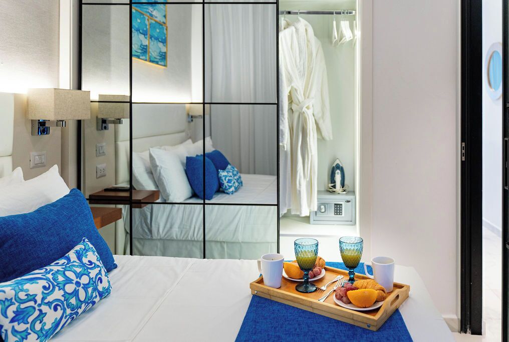 Une chambre d’hôtel avec un lit, un plateau de petit-déjeuner comprenant du jus d’orange et des croissants, et un miroir qui reflète une salle de bain avec un peignoir et une douche.