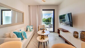 A modern hotel room with a sofa, two round tables, a TV, and a balcony with a view.