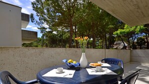 A round table with chairs, a vase with flowers, and a plate of food on an outdoor patio with a concrete wall and trees in the background. - Parco Hemingway by Interhome (Lignano Pineta)