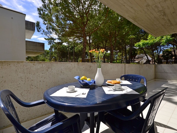 A round table with chairs, a vase with flowers, and a plate of food on an outdoor patio with a concrete wall and trees in the background.