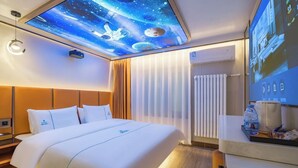 Habitación de hotel con una cama grande, un proyector en el techo que muestra una escena espacial, una mesita de noche con una lámpara y un panel de control en la pared.