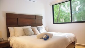 Una cama bien hecha con cabecera de madera, una ventana con vistas a árboles y una mesita de noche con una lámpara.