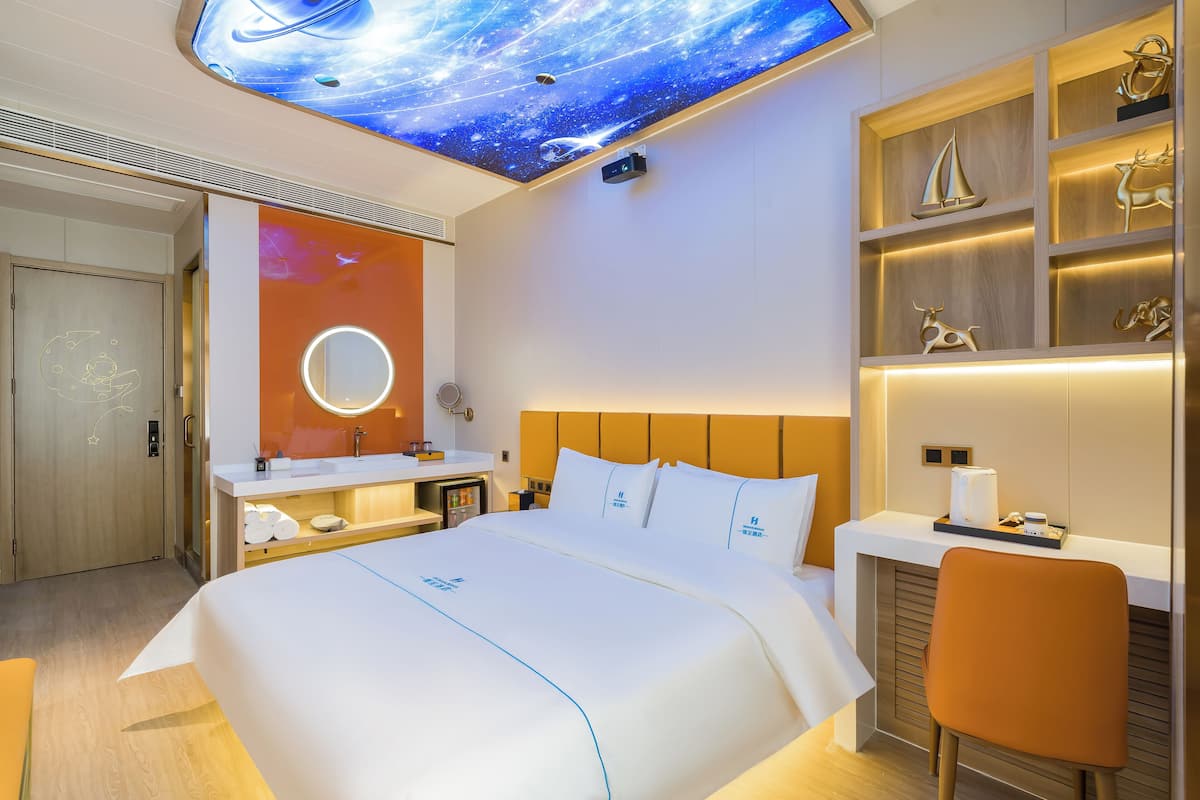 A modern hotel room with a galaxy-themed ceiling, a large bed, a bedside table, a chair, and a built-in shelf with decorative items.