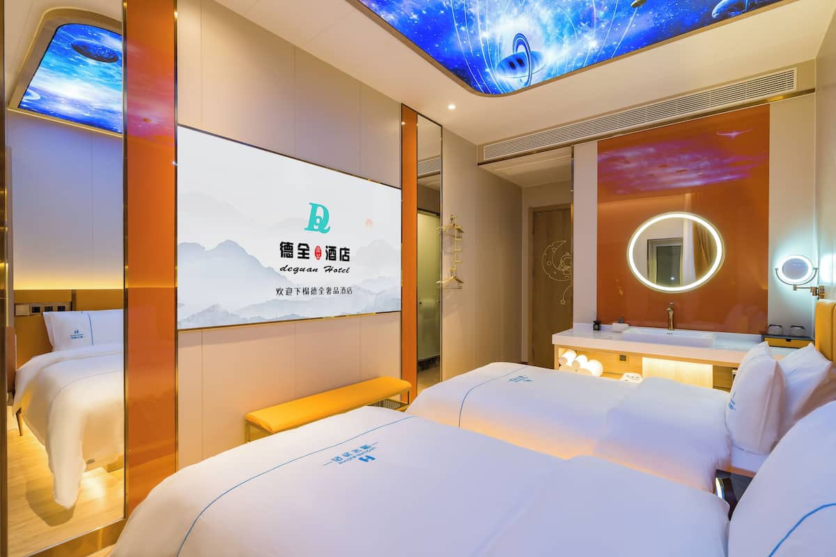 A hotel room with two beds, a large digital screen displaying the hotel name, a bathroom with a round mirror, and a night sky-themed ceiling.