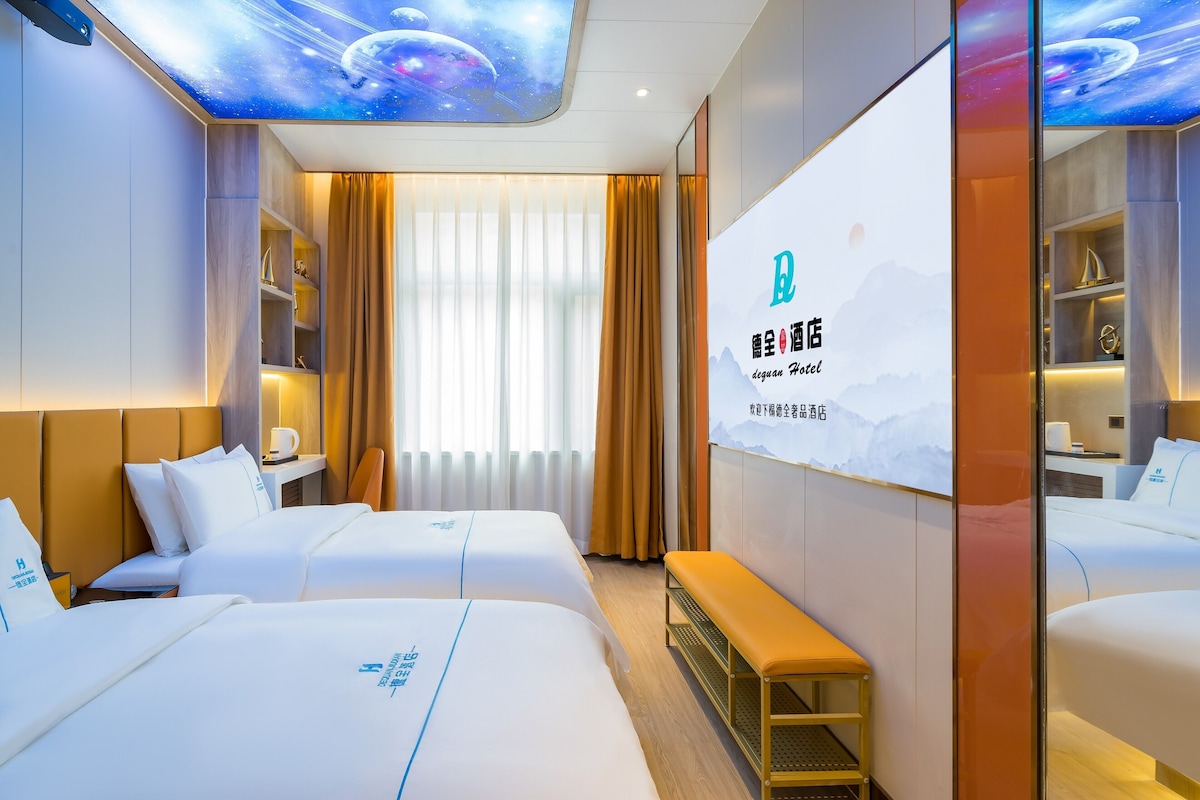 A hotel room with two beds, a bench, a desk, and a large screen displaying the hotel name.