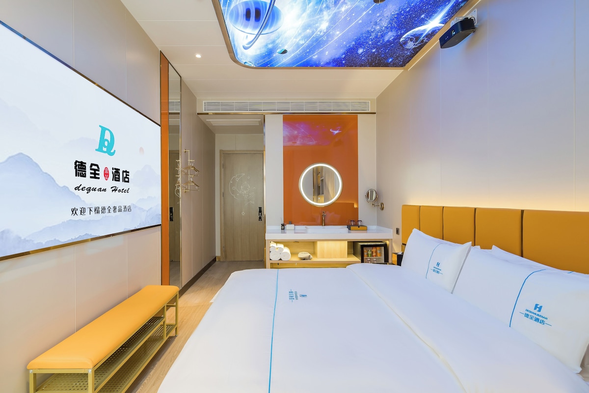 A hotel room with a large bed, a bench, a wall-mounted TV, and a bathroom with a mirror and sink.