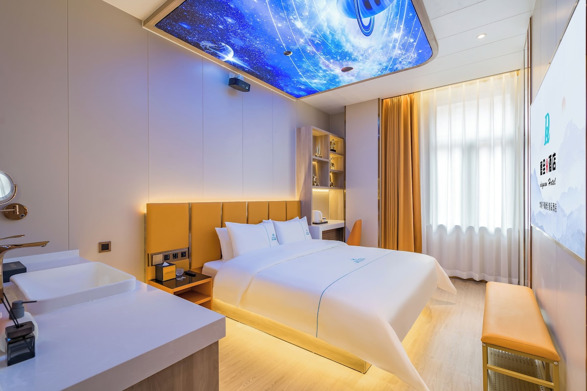 A modern hotel room with a large bed, a bedside table, a built-in shelf, a ceiling projector displaying a starry sky, and a large window with curtains.
