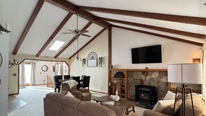 A spacious living room with a high ceiling, exposed wooden beams, a fireplace, a television, and a dining area with a table and chairs. - Live & Love Life on the Lake at Sunset Peak! (Howe)