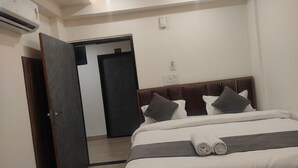 A hotel room with a bed, a wooden headboard, a wall-mounted air conditioner, a door, and a light switch. - Hotel Abhinandan (Ayodhya)
