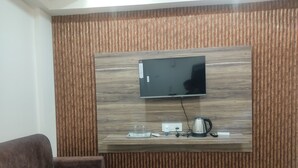 A modern living room with a flat-screen TV mounted on a wooden wall, a sofa, a coffee table, and a kettle on the counter. - Hotel Abhinandan (Ayodhya)