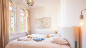 Une chambre bien éclairée, avec un grand lit, une table de chevet sur laquelle se trouve une bouteille de champagne, et un luminaire fixé au mur.