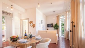 Un coin repas avec une table ronde, des chaises blanches et un saladier rempli de fruits. On aperçoit un canapé et un téléviseur au fond.