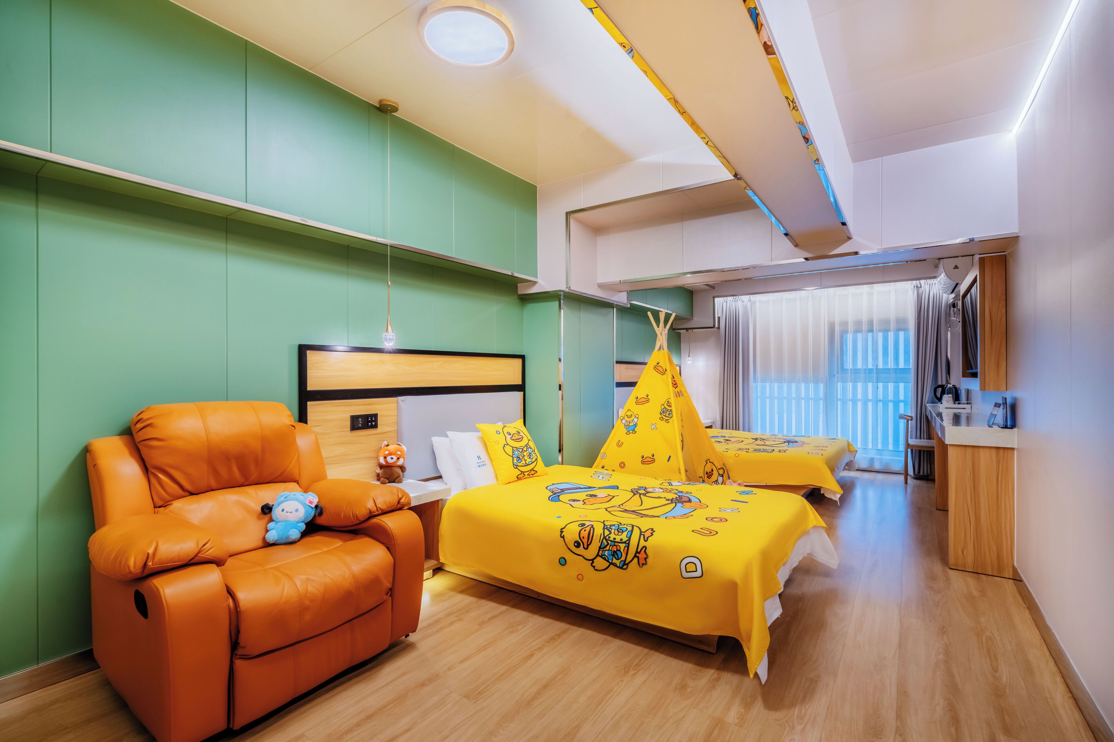 A hotel room with a sofa, two beds with yellow duck-themed bedding, a wooden desk, and a window with curtains.