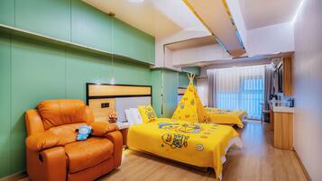 A hotel room with a sofa, two beds with yellow duck-themed bedding, a wooden desk, and a window with curtains.
