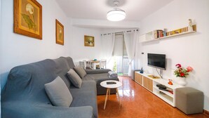 A living room with a grey sofa, a small round table, a TV on a wooden stand, and a bookshelf with decorative items. - Fuente de Piedra Apartment (Torre Del Mar)
