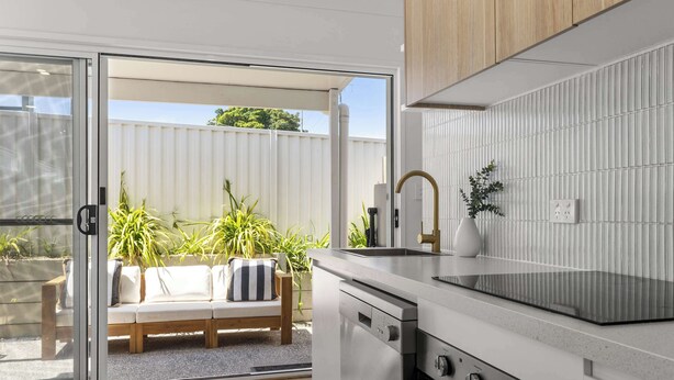 Une cuisine moderne dotée d’un four encastré et d’une porte-fenêtre coulissante donnant sur un espace extérieur aménagé avec un canapé blanc et des plantes.