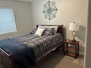 A bedroom with a bed, a bedside table with a lamp, a decorative wall piece, and a window with blinds. - Charming 3-Bedroom Retreat in Hazel Dell, WA – Your Perfect Getaway! (Vancouver)