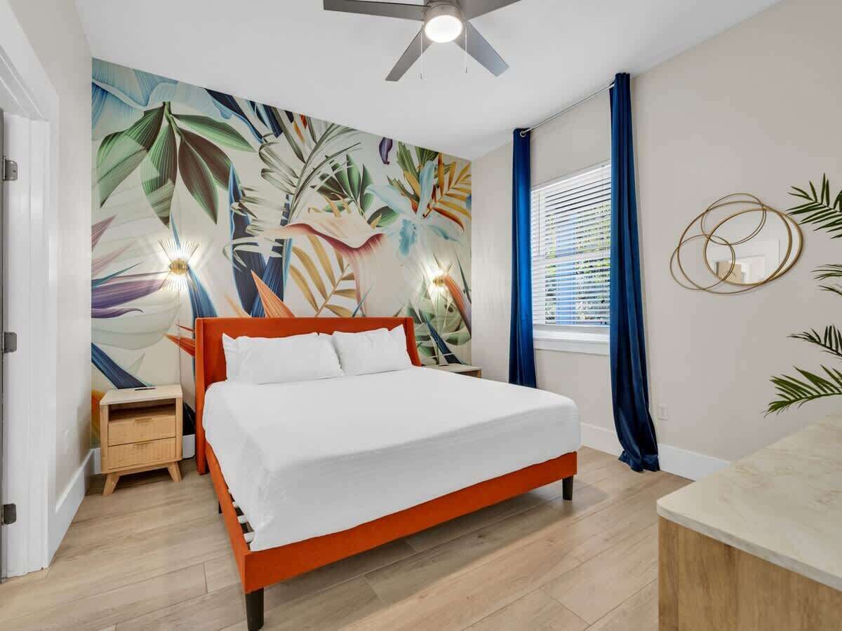 Un dormitorio con cama, mesita de noche, ventilador de techo, ventana con cortinas y un mural en la pared.