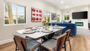 A modern dining area with a white table set for six, surrounded by dark chairs. There are large windows on one side, a blue sofa in the background, and a flat-screen TV mounted on the wall.